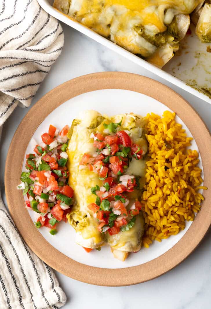 Top down view white plate with a serving of veggie enchiladas with green sauce and pico de gallo, nestled between a serving of pico de gallo and spanish rice.